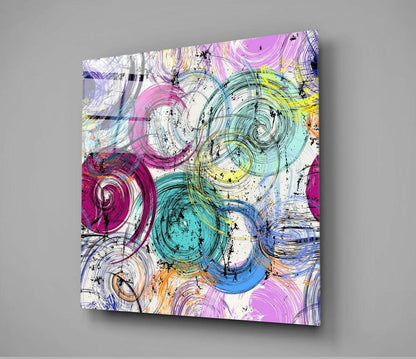 Colorful glass wall art panel featuring overlapping spiral and circular designs in magenta, teal, yellow, and blue with black line work, displayed at an angle against gray background.