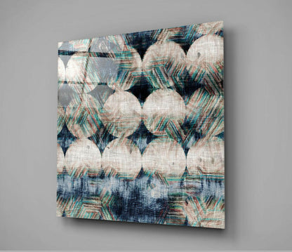 Square glass wall art panel displaying overlapping cream and teal patterned fabric design with dark blue accents, mounted at angle against gray background.