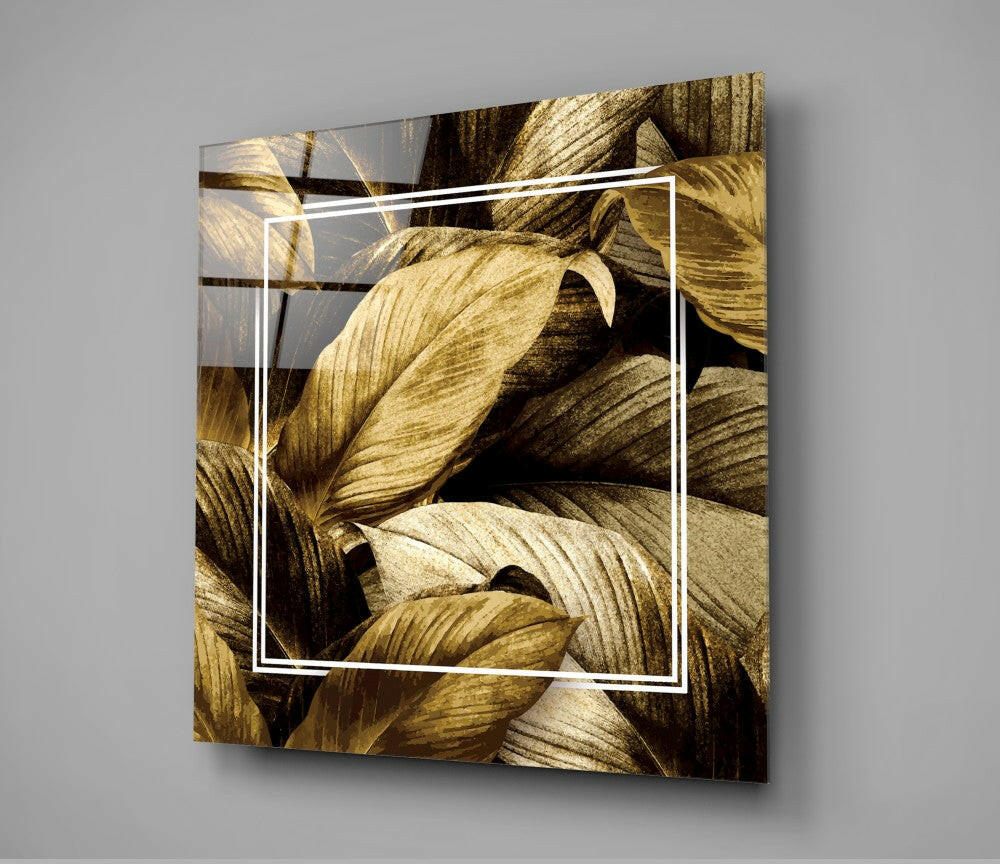 Gold leaf glass wall art displaying layered golden dried leaves with a white rectangular frame overlay, mounted at an angle against gray background.