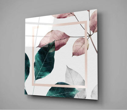 Angled view of square glass wall art with watercolor leaves in mauve, teal, and gray tones framed by a pale pink border, displayed against gray background.