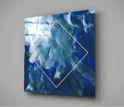 Square glass wall art with rotated diamond outline displaying blue and white brushstroke abstract painting, mounted on gray wall.