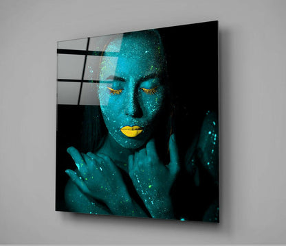 Angled view of glass wall art displaying a woman's face in turquoise with gold eyelashes and lips against black background.