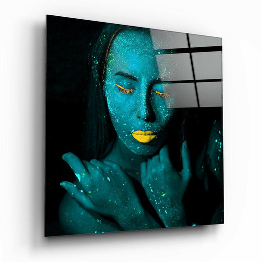 A glass wall art panel displaying a woman's face painted turquoise with gold accents, shown at an angled perspective against a white background.