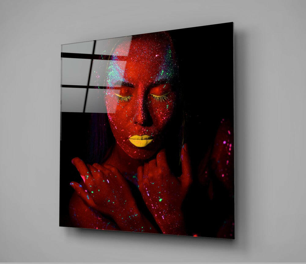 Ruby Woman Glass Wall Art displayed at an angle, showing a woman's face covered in red glitter with yellow lips and closed eyes, mounted on gray wall.