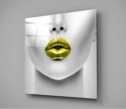 Modern glass wall art displaying a minimalist white sculptural face with striking gold metallic lips, mounted at an angle against a gray background.