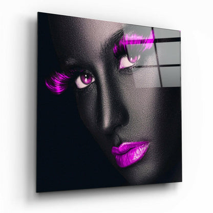 Angled view of glass wall art displaying a grayscale face with vibrant magenta eyes, lashes, and lips against a dark background.