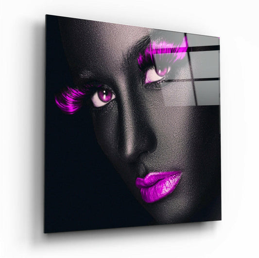 Angled view of glass wall art displaying a grayscale face with vibrant magenta eyes, lashes, and lips against a dark background.
