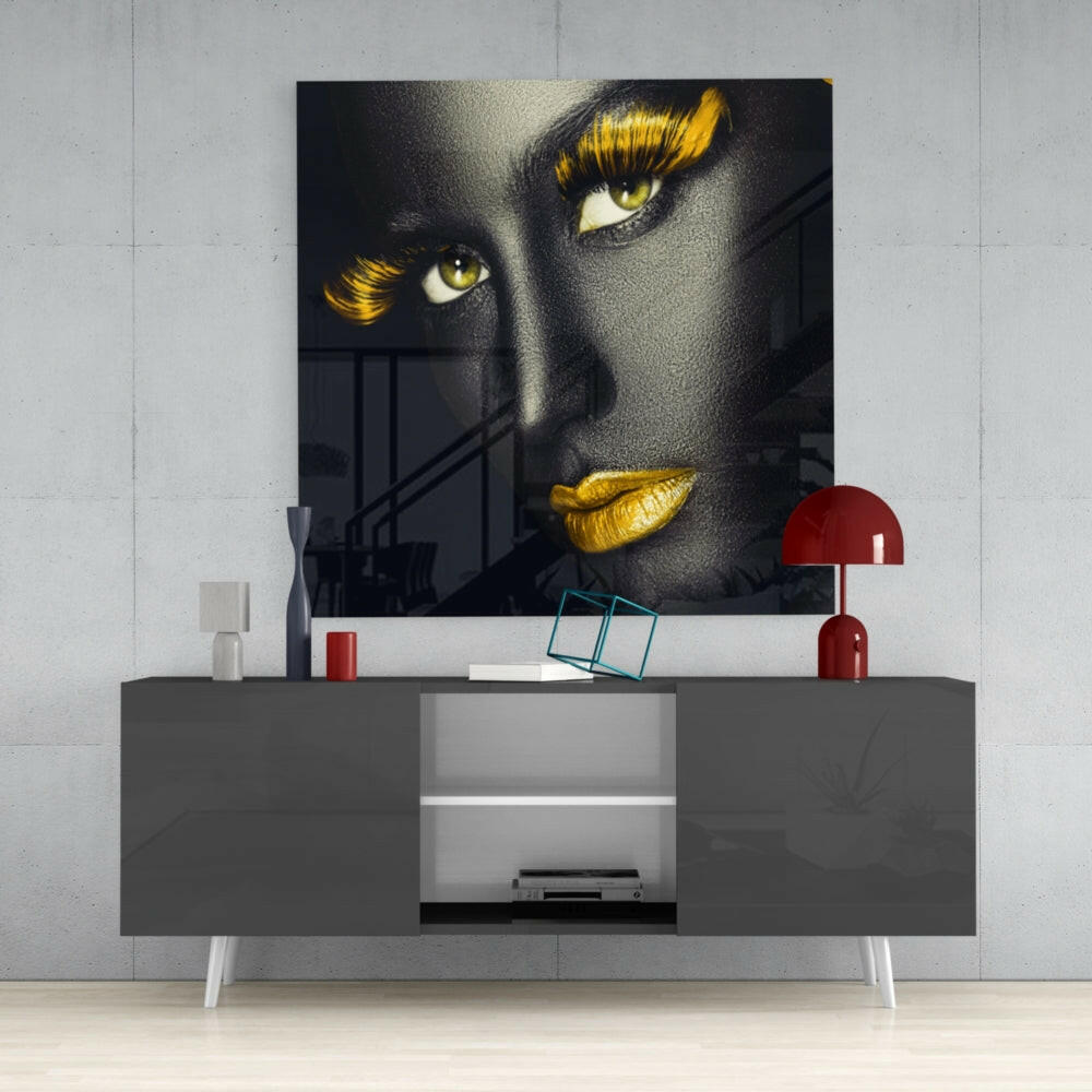Glass wall art featuring a woman's face with black skin, golden eyeshadow and lips, displayed above a gray media console in a modern interior.