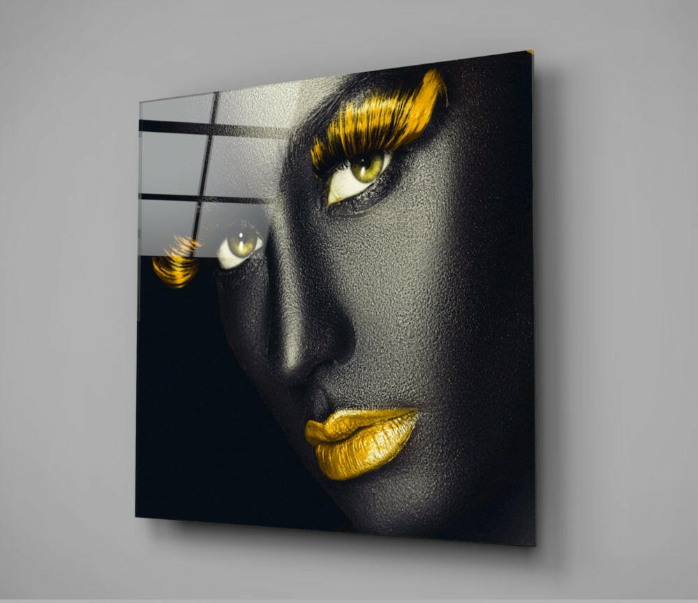 Modern glass wall art displaying a portrait with dramatic black skin, golden yellow eyeshadow, and gold lips, mounted at an angle against a gray wall.