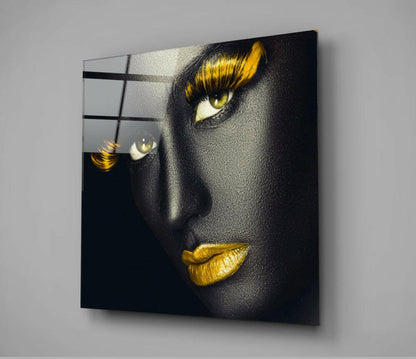 Modern glass wall art displaying a portrait with dramatic black skin, golden yellow eyeshadow, and gold lips, mounted at an angle against a gray wall.