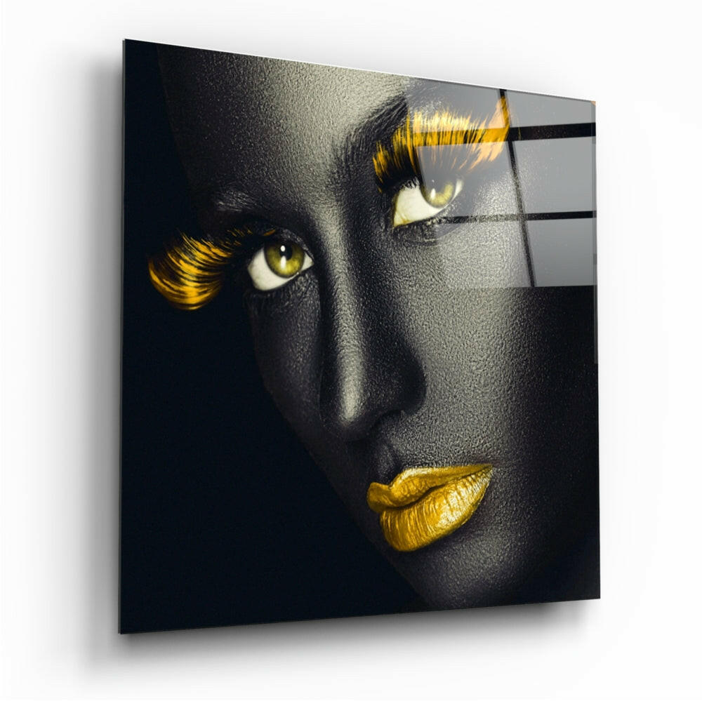 Glass wall art mounted at an angle displaying a face with black skin, golden eyes, yellow lashes, and gold lips against a dark background.