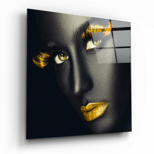 Glass wall art mounted at an angle displaying a face with black skin, golden eyes, yellow lashes, and gold lips against a dark background.