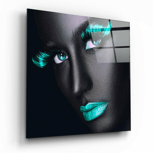 Angled view of square glass wall art featuring a woman's face with turquoise glowing eyes, lashes, and lips against black background.