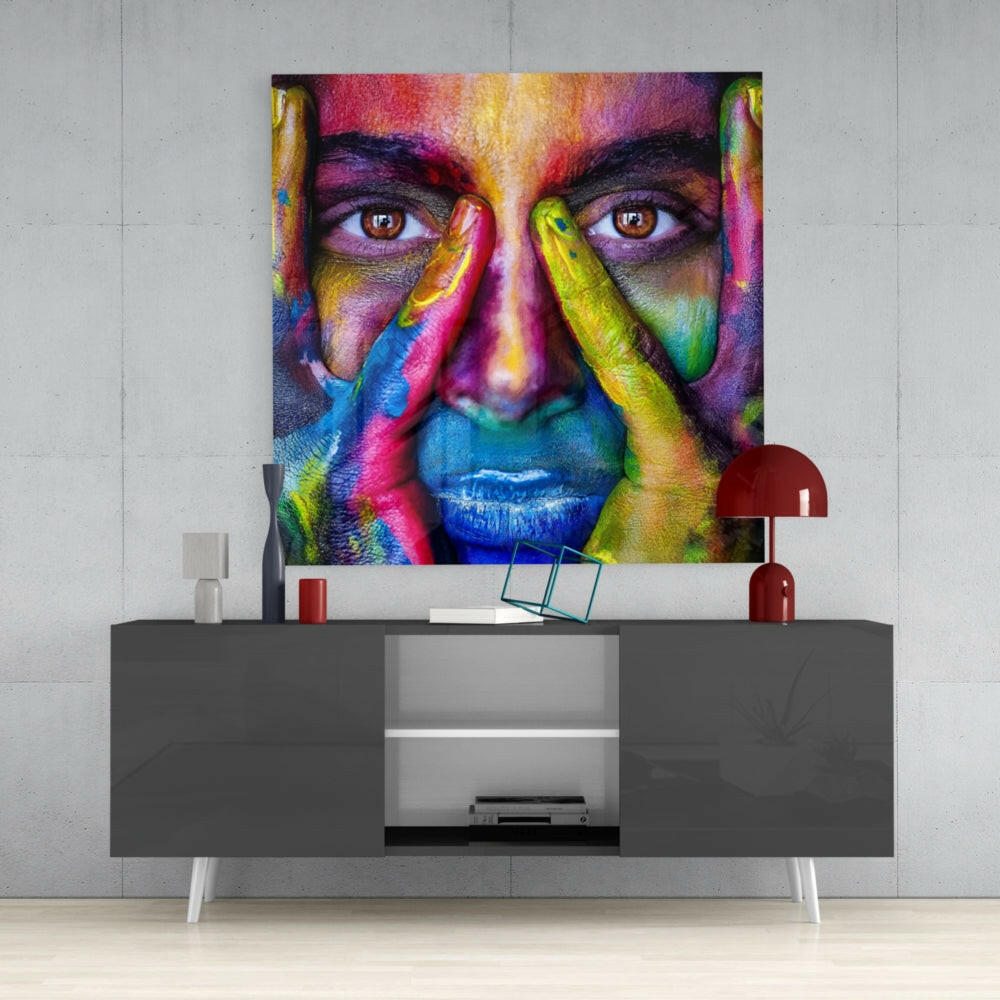 Colorful glass wall art featuring a close-up portrait with rainbow-painted face and hands displayed above a gray media console.