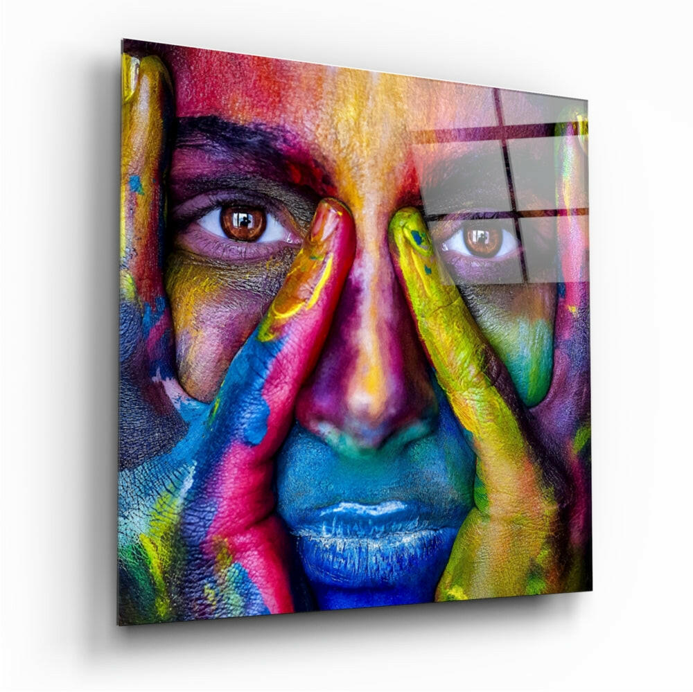 Colorful glass wall art displaying a close-up portrait of a face painted in vibrant rainbow hues with hands framing the face, shown at an angled perspective.