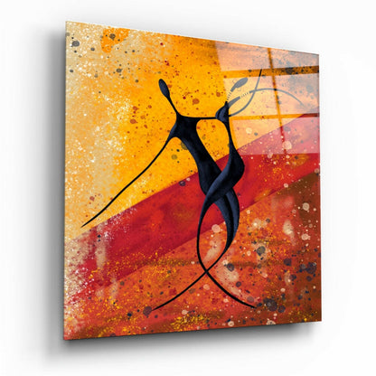 Glass wall art panel with black silhouette figures dancing against warm gradient background of yellow, orange, and red with spattered texture effects.