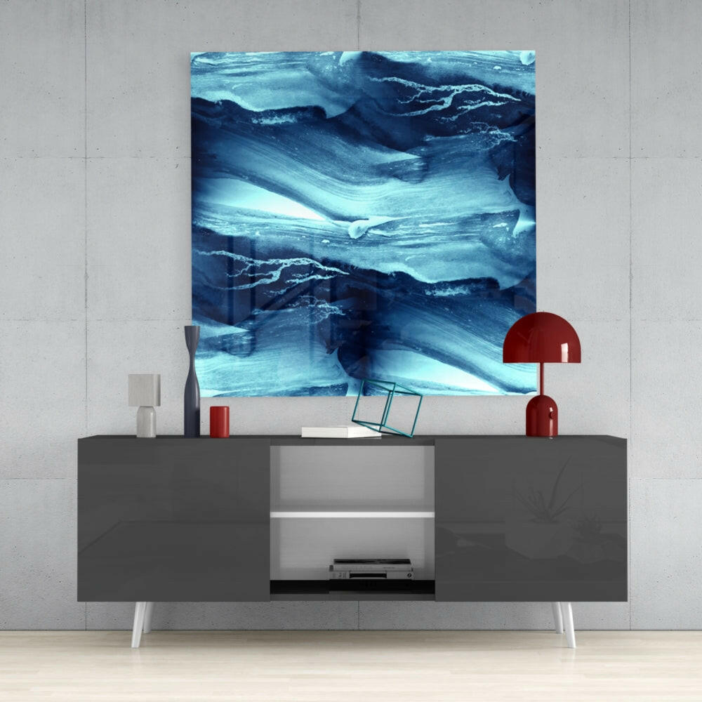 Abstract Waves Glass Wall Art in shades of blue and white displayed above a gray media console with red lamp and accessories.