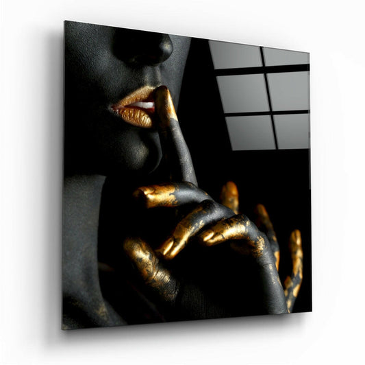 Close-up portrait of a figure with dark skin and gold-painted lips holding a finger to mouth in a "shush" gesture against a black background.