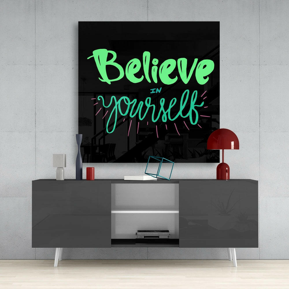Neon glass wall art displaying "Believe in Yourself" in green and cyan lettering mounted above a gray media console in a modern interior setting.