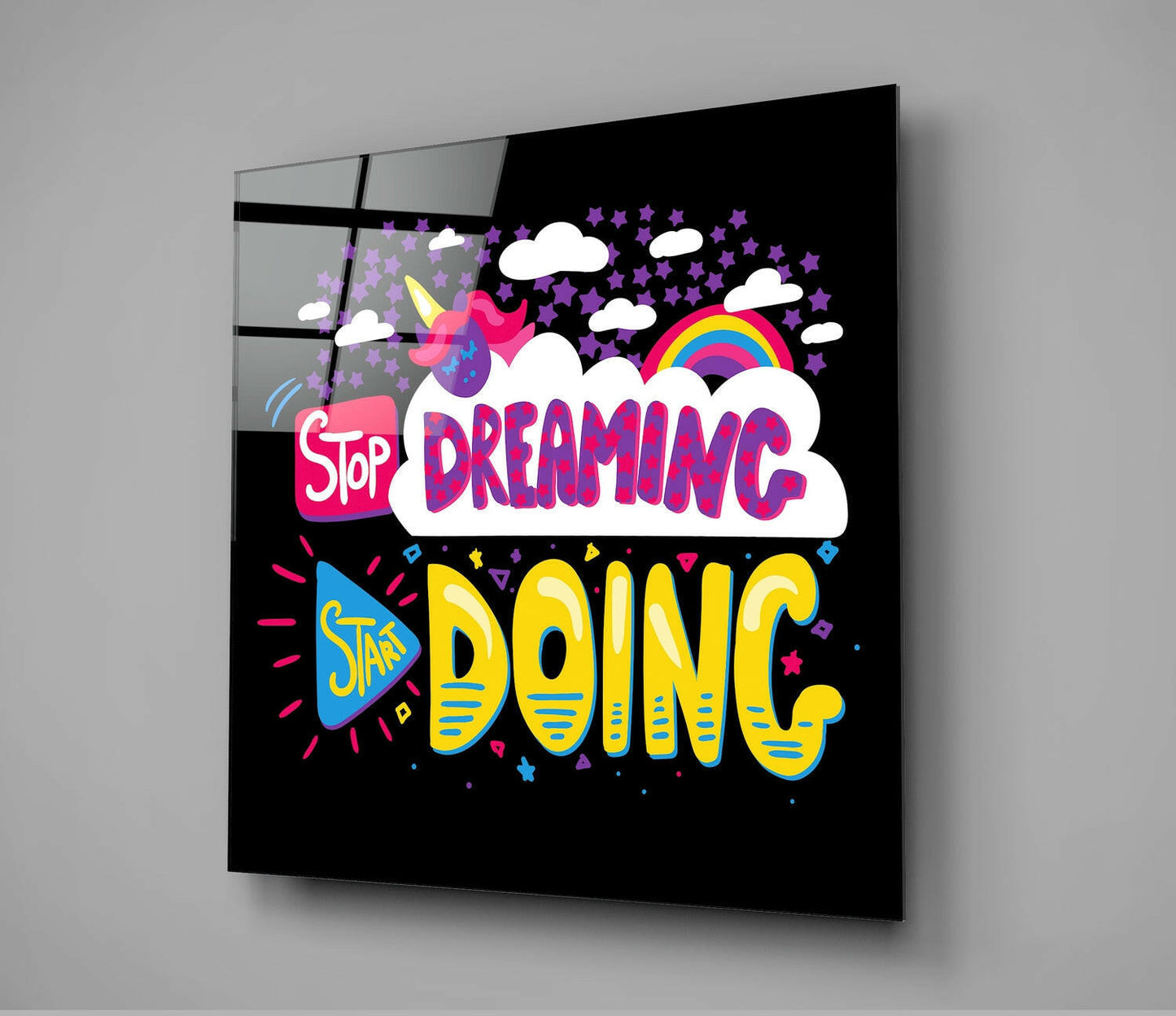 Angled view of black glass wall art panel with colorful "Stop Dreaming Start Doing" text, unicorn, rainbow, clouds, and stars on dark background.