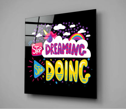 Angled view of black glass wall art panel with colorful "Stop Dreaming Start Doing" text, unicorn, rainbow, clouds, and stars on dark background.