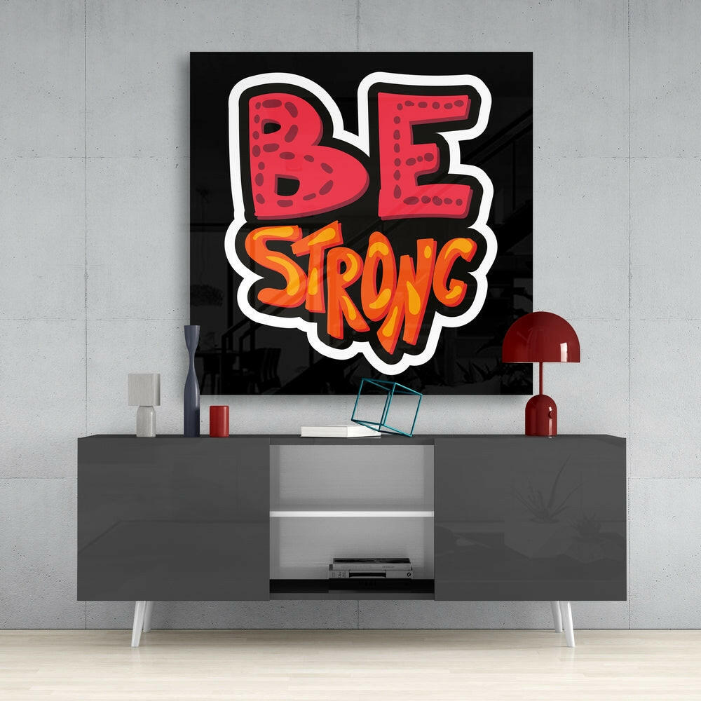 Black glass wall art displaying "BE STRONG" in red and orange bubble letters mounted above gray media console in modern interior.