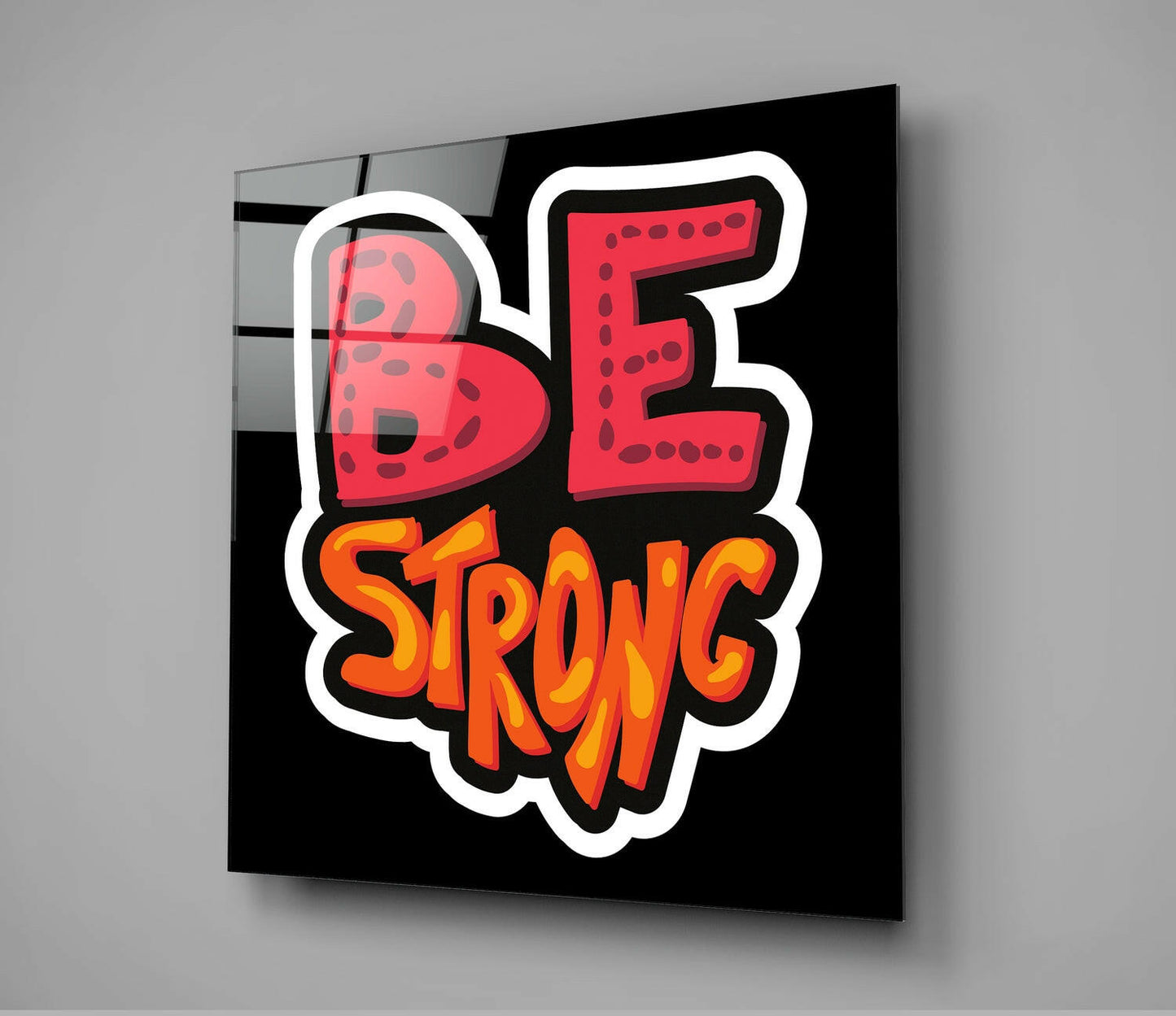 Black glass wall art panel displaying red "BE" and orange "STRONG" text with white outline, photographed at an angle against gray background.
