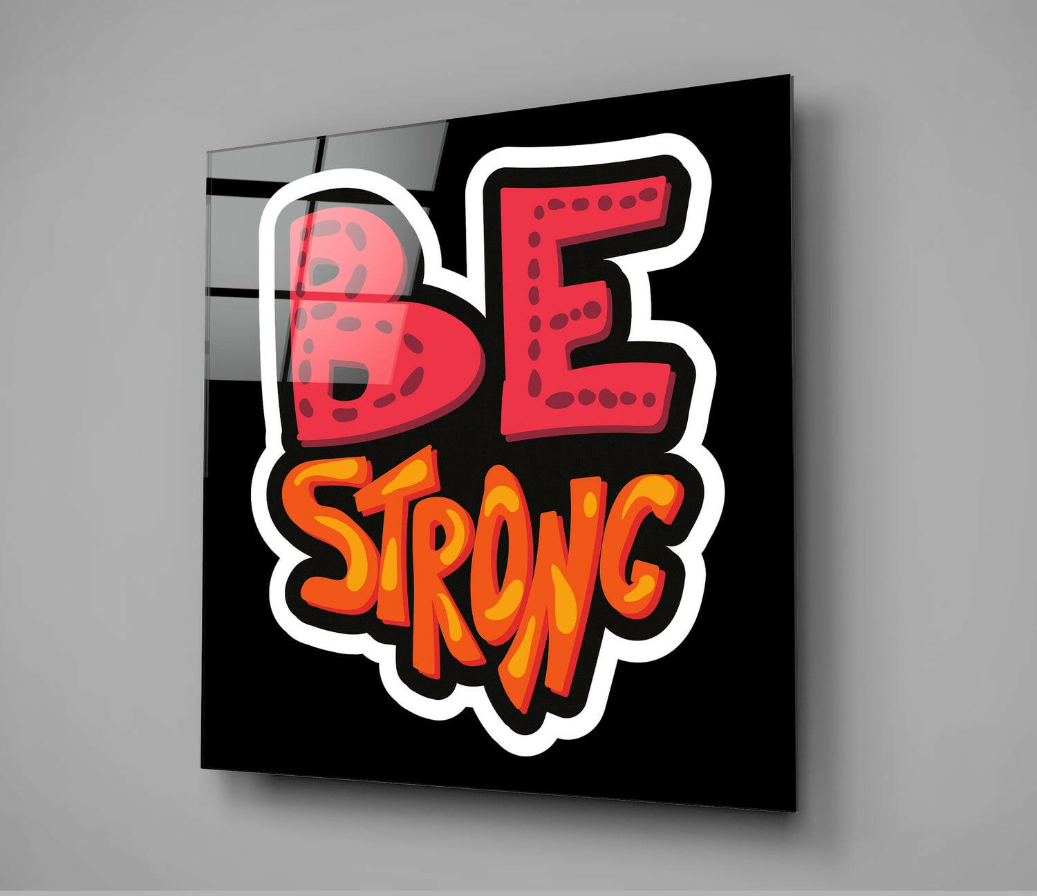 Black glass wall art panel displaying red "BE" and orange "STRONG" text with white outline, photographed at an angle against gray background.
