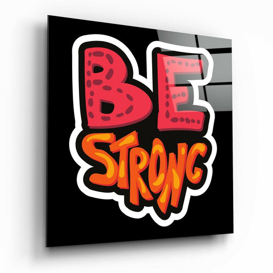 Glass wall art panel displayed at an angle showing red "BE" letters and orange "STRONG" text with white border on black background.