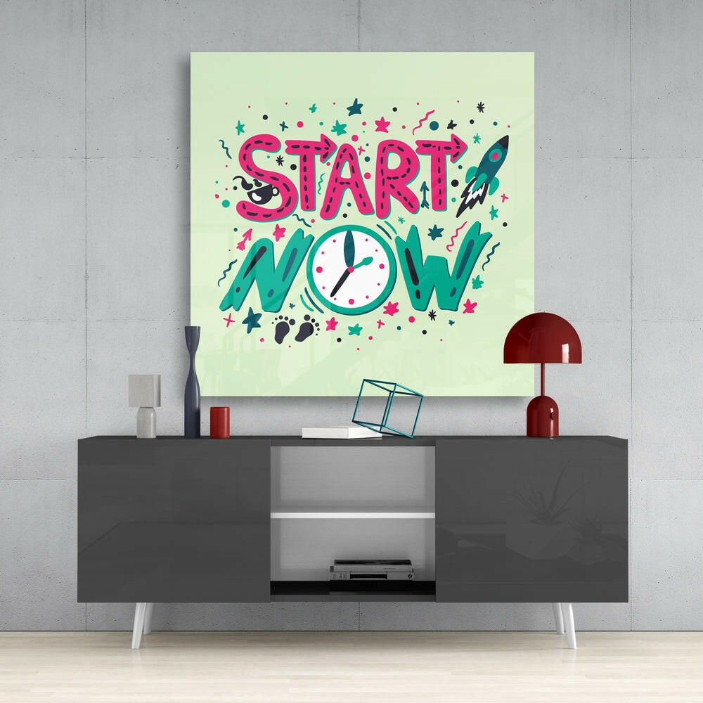 Lime green glass wall art displaying "START NOW" in pink and teal lettering with decorative stars, clock, and rocket motifs, mounted above a gray media console.
