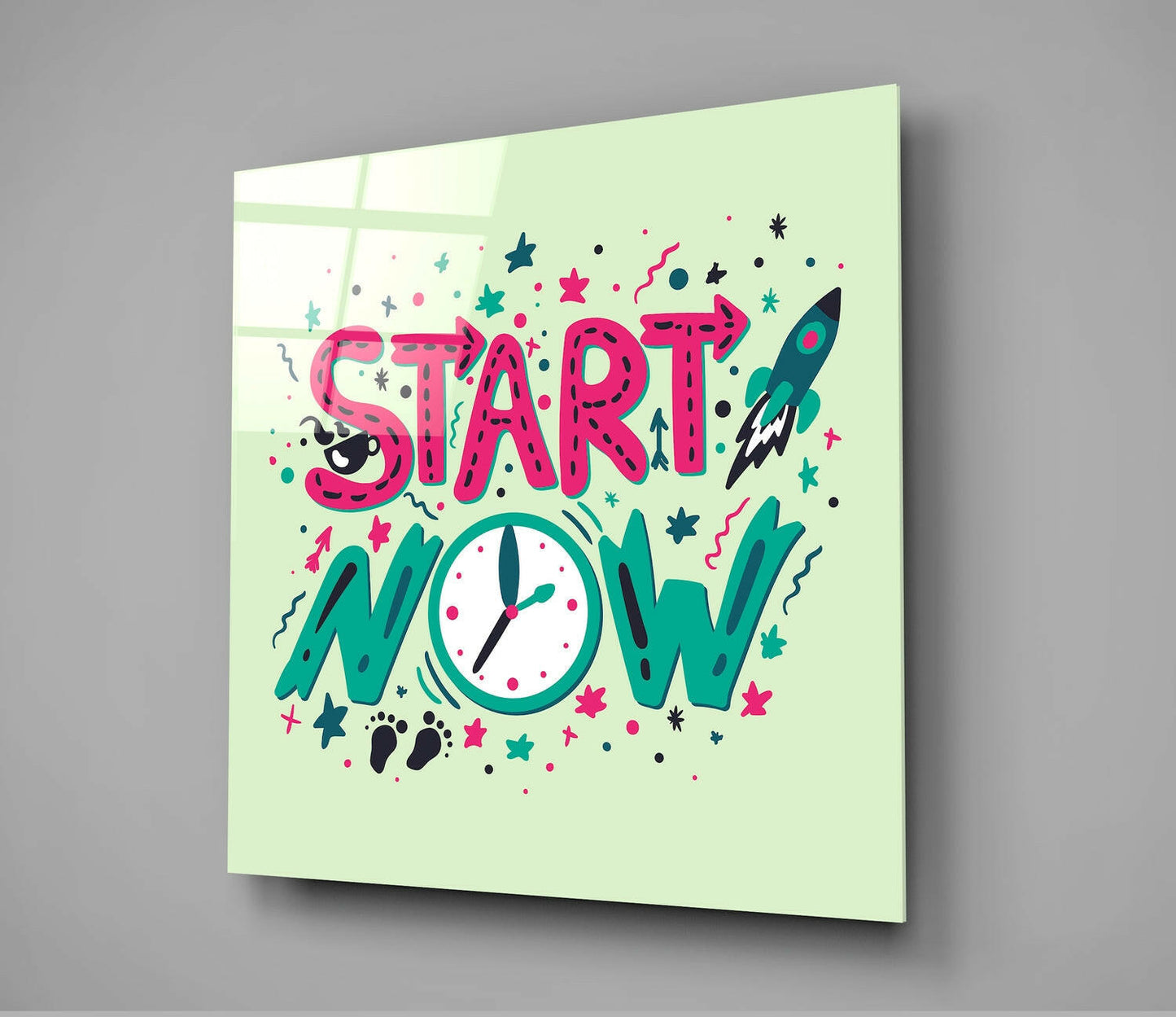 Angled view of a lime green glass wall panel with pink "START NOW" text, teal "NOW" letters, a clock face, rocket, stars, and decorative elements in pink and teal colors.