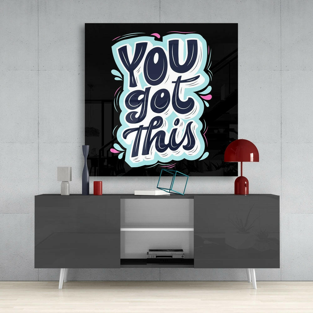 Neon-style glass wall art displaying "You Got This" in cyan, navy, and pink lettering mounted above a gray media console in a modern interior setting.