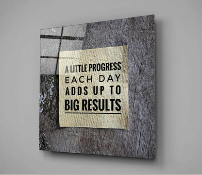 Glass wall art with cream-colored center panel displaying black text "A LITTLE PROGRESS EACH DAY ADDS UP TO BIG RESULTS" mounted on textured gray wood background.