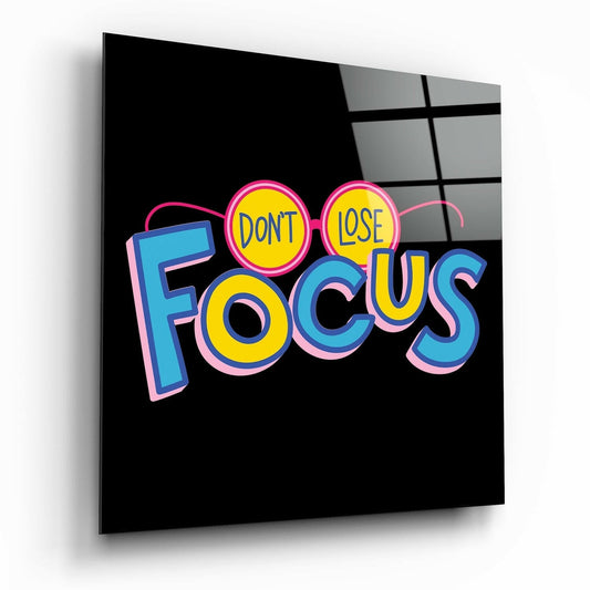 Angled view of glass wall art displaying colorful neon-style text reading "DONT LOSE FOCUS" with pink, blue, and yellow lettering on black background.