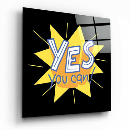 Glass wall art panel displayed at an angle showing a yellow starburst with "YES you can!" text in blue and orange lettering against a black background.