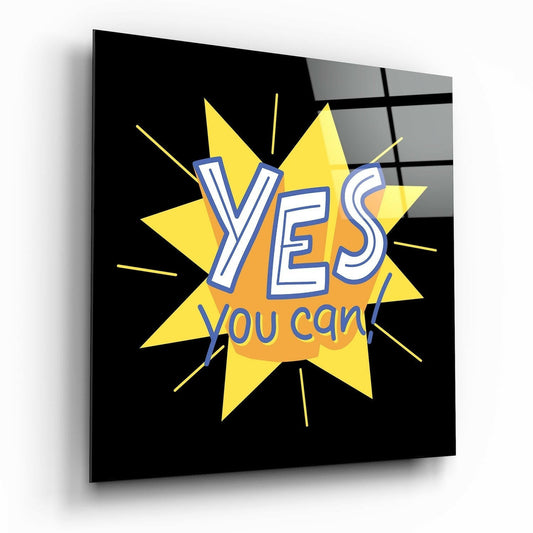 Glass wall art panel displayed at an angle showing a yellow starburst with "YES you can!" text in blue and orange lettering against a black background.