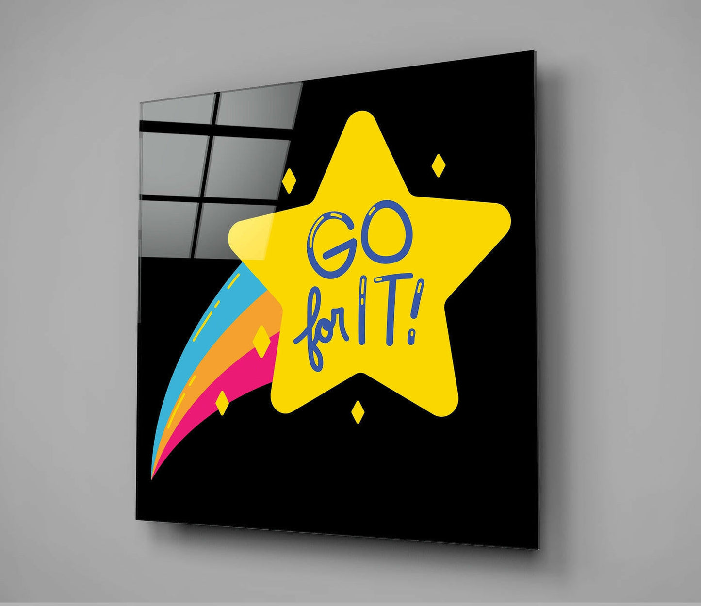 Glass wall art panel displaying a bright yellow star with "GO for IT" text in blue, colorful comet trail, and small diamond shapes on black background, shown at an angle against gray wall.