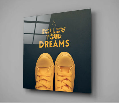 Glass wall art mounted on gray wall showing yellow sneakers and gold text "Follow Your Dreams" on dark teal background.