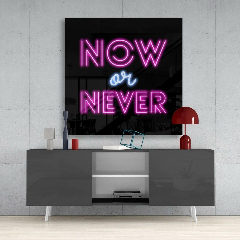 Neon glass wall art displaying "NOW or NEVER" in pink and blue lettering, mounted above a gray modern console table with red lamp and accessories.