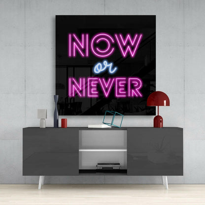 Neon glass wall art displaying "NOW or NEVER" in pink and blue lettering, mounted above a gray modern console table with red lamp and accessories.