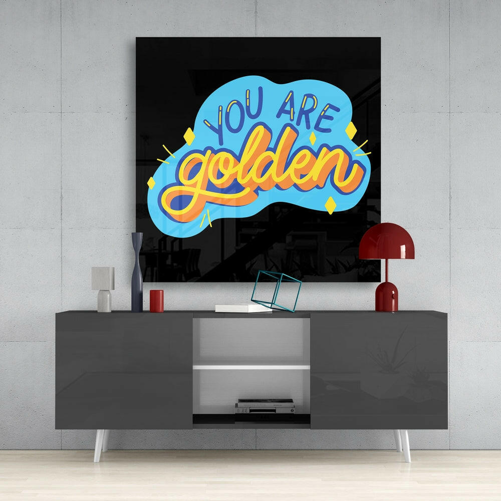 Neon-style glass wall art displaying "You Are Golden" in blue and yellow lettering, mounted above a gray media console in a modern interior setting.