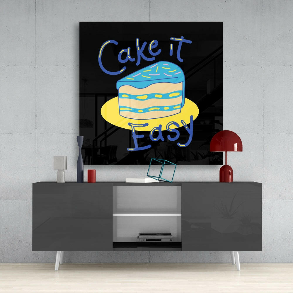 Neon-style glass wall art displaying "Cake it Easy" text in blue with an illustrated layered cake in blue and yellow, mounted above a gray media console.