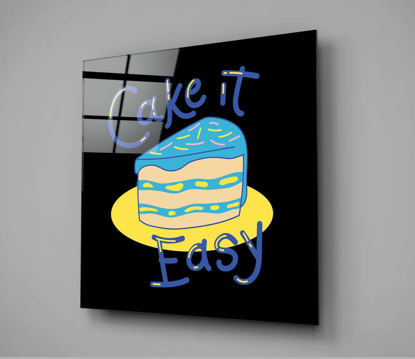 Angled view of black glass wall art with neon blue "Cake It Easy" text and colorful illustrated cake on yellow plate.
