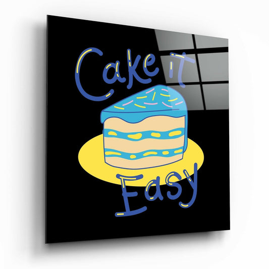 Angled perspective view of black glass wall art featuring a blue and yellow layered cake illustration with "Cake It Easy" text in blue lettering.