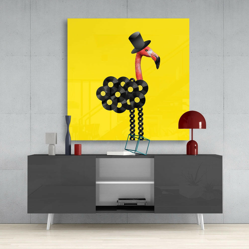 Bright yellow glass wall art featuring a flamingo wearing a black top hat with black patterned body, displayed above a gray media console.
