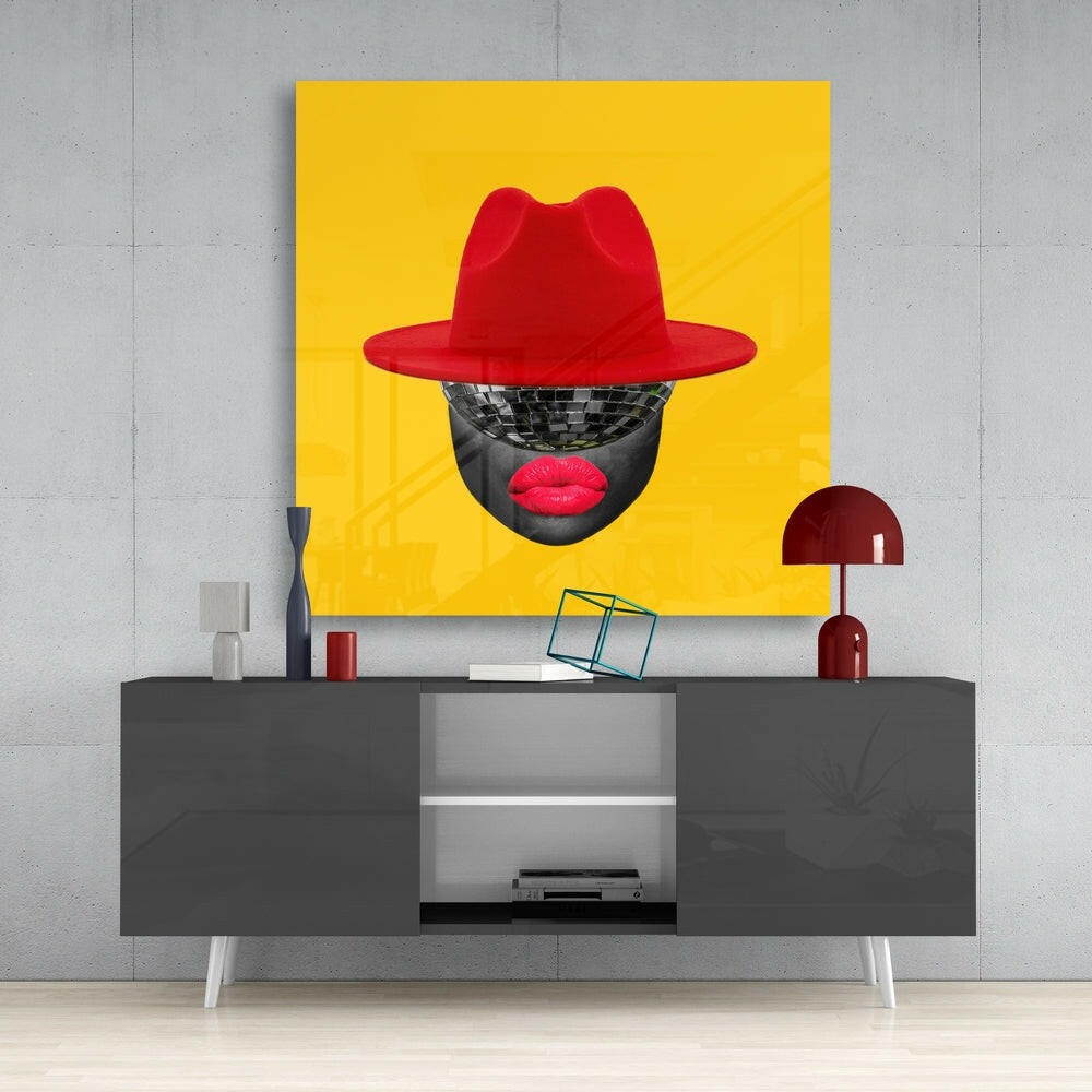 Bold yellow glass wall art with red fedora hat and disco ball face against grey concrete wall above modern grey console table.