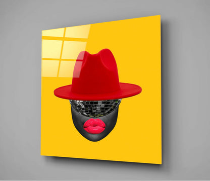 Angled view of bright yellow glass wall art featuring a stylized face with disco ball eyes, red lips, and red fedora hat.