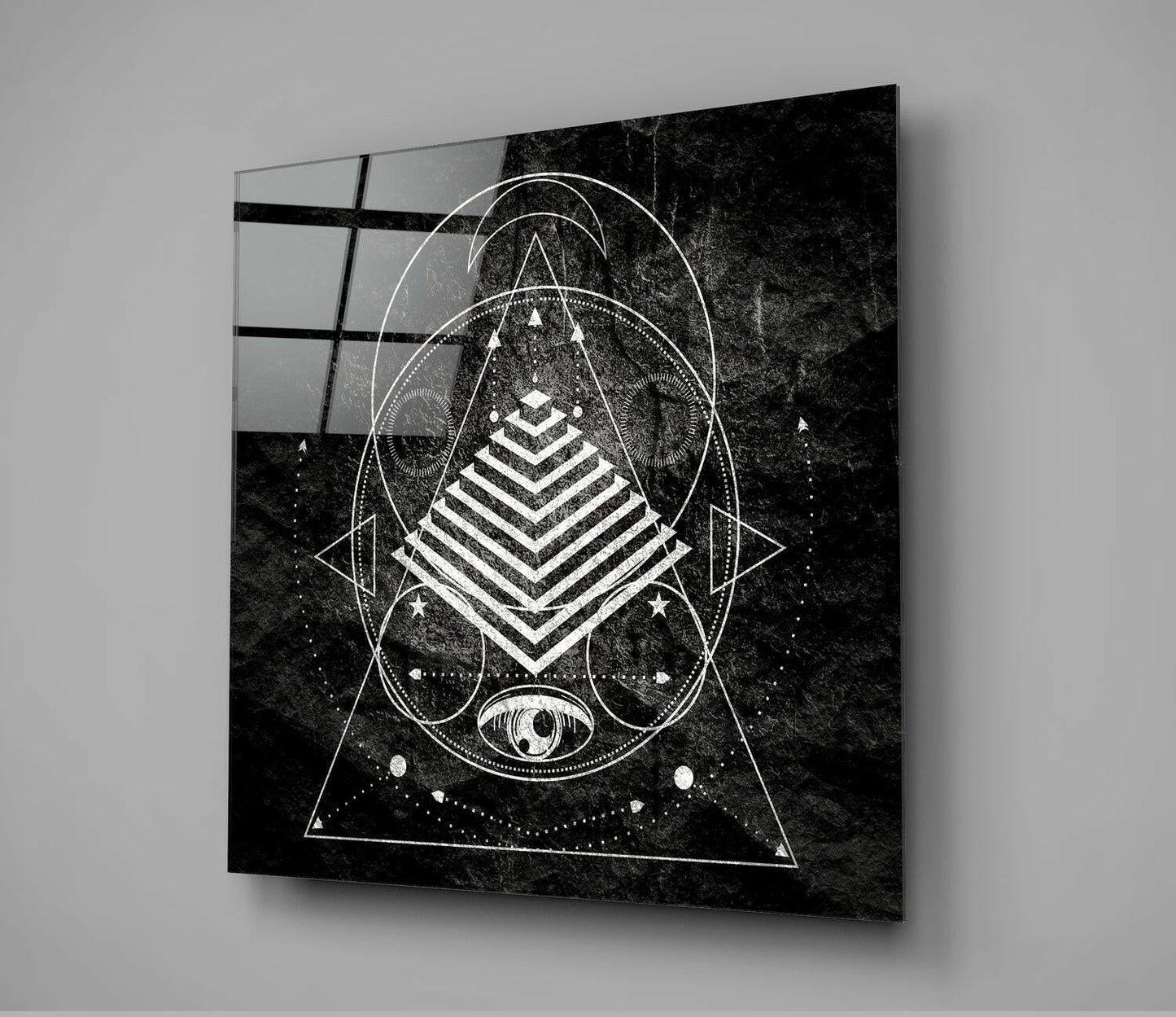 Black and white geometric sacred design printed on glass panel mounted on gray wall, displaying nested triangles, pyramid, circles, and celestial eye motif.