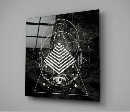 Black and white geometric sacred design printed on glass panel mounted on gray wall, displaying nested triangles, pyramid, circles, and celestial eye motif.