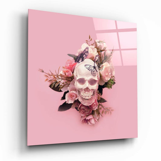 Glass wall art panel displaying a skull adorned with pink roses and butterflies against a pink background, viewed at an angle showing the glass surface and shadow.